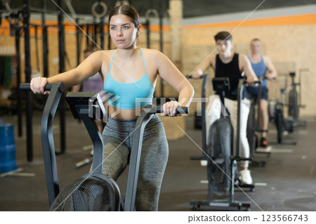 Young dedicated woman training on exercise bike near other people in gym Young dedicated woman training on exercise bike near other people in gym 123566743