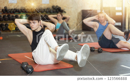 Concentrated young man doing abdominal exercise on yoga mat during group workout in gym. CrossFit healthy concept Concentrated young man doing abdominal exercise on yoga mat during group workout in gym. CrossFit healthy concept 123566888