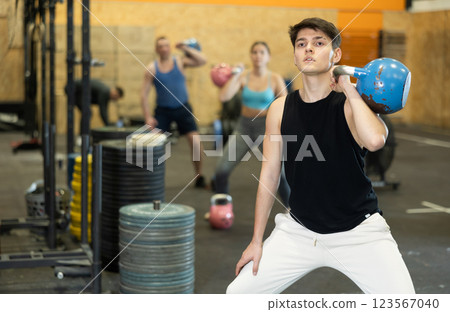 Active young man in sportswear exercising CrossFit with kettlebell while training with group in fitness center 123567040