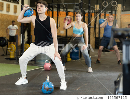 Sportive strong young male in activewear swinging kettlebell during group exercise class in CrossFit gym indoors 123567115