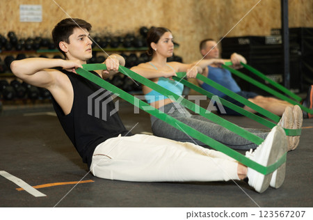 Motivated young man practicing exercises with stretch rope sitting near other people in gym 123567207