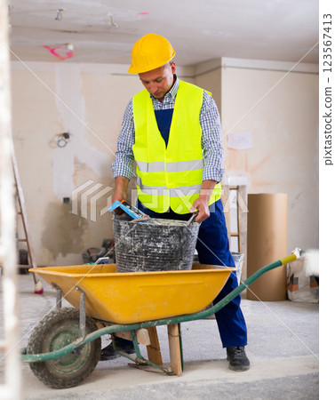 Worker in yellow vest and a protective helmet transports painting tools in wheelbarrow 123567413