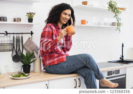 Smiling African American woman sitting on kitchen counter talking on phone holding coffee mug. Relaxed, casual atmosphere within modern home interior. Smiling African American woman sitting on kitchen counter talking on phone holding coffee mug. Relaxed, casual atmosphere within modern home interior. 123568356