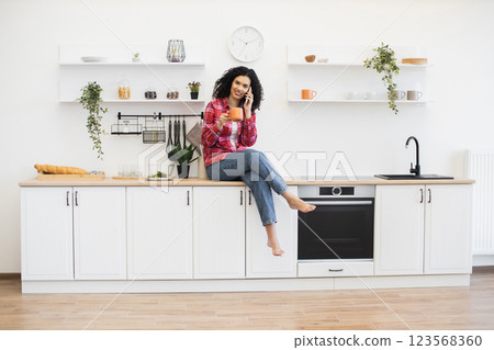 Young African American woman enjoying casual moment in bright kitchen holding coffee cup while communicating via mobile phone, wearing casual attire, sitting on kitchen countertop. Young African American woman enjoying casual moment in bright kitchen holding coffee cup while communicating via mobile phone, wearing casual attire, sitting on kitchen countertop. 123568360