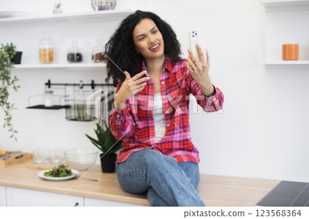 African young woman in casual outfit video chatting with cheerful expression sitting in bright kitchen. African young woman in casual outfit video chatting with cheerful expression sitting in bright kitchen. 123568364