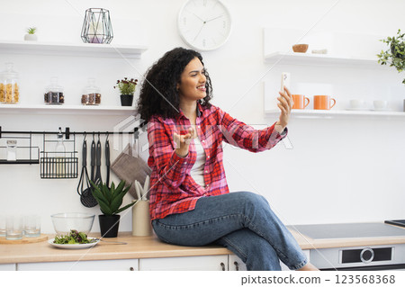 Smiling African woman sitting on kitchen counter holding smartphone taking selfie, expressing joy and relaxation, surrounded by kitchenware, plants, and a healthy salad. Smiling African woman sitting on kitchen counter holding smartphone taking selfie, expressing joy and relaxation, surrounded by kitchenware, plants, and a healthy salad. 123568368
