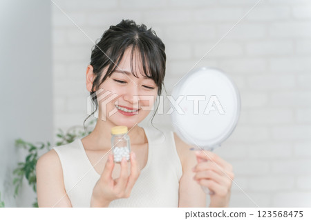 Smiling Asian woman holding a bottle of medicine/supplements while looking in the mirror 123568475
