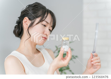 Asian woman with vitamins and supplements suffering from rough skin and acne looking in the mirror 123568476