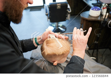 Man barber cutting little boy's hair using comb and scissors. Child getting haircut from adult male, likely barber. Professional hairdresser and cute client at modern barbershop. Man barber cutting little boy's hair using comb and scissors. Child getting haircut from adult male, likely barber. Professional hairdresser and cute client at modern barbershop. 123568844