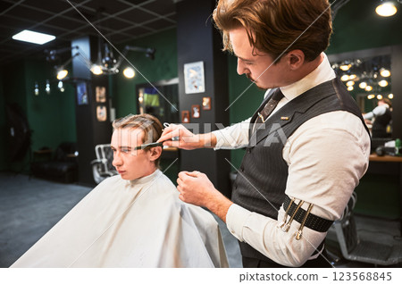 Barber giving haircut to young man in barbershop. Professional barber well-dressed in vest and white shirt with rolled-up sleeves, concentrating on cutting the hair, using comb and scissors. Barber giving haircut to young man in barbershop. Professional barber well-dressed in vest and white shirt with rolled-up sleeves, concentrating on cutting the hair, using comb and scissors. 123568845