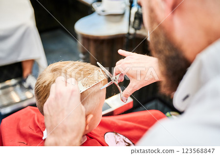 Man barber cutting little boy's hair using comb and scissors. Child getting haircut from adult male, likely barber. Professional hairdresser and cute client at modern barbershop. 123568847
