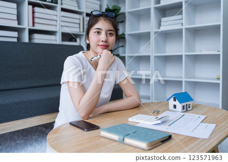 Thoughtful young woman planning home project while sitting at a table with a miniature house model and notebooks 123571963