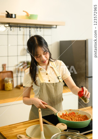 Smiling young woman adding spices into a simmering pan of tomato based spaghetti sauce Smiling young woman adding spices into a simmering pan of tomato based spaghetti sauce 123572965