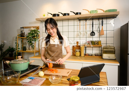 Cheerful young woman wearing a apron slicing carrot while preparing ingredients for homemade spaghetti dish 123572987