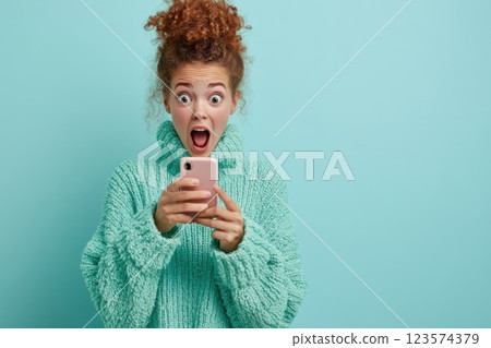 Young woman shows startled expression while dropping her phone against pale blue background 123574379