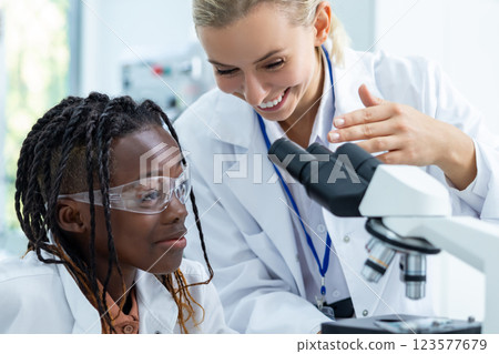 Blonde teacher in white lab explaining how to work with microscope to a student 123577679