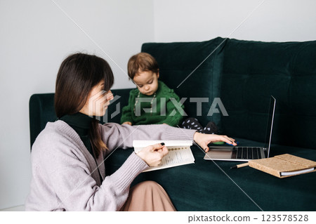 Mother working remotely with toddler on couch. Remote work challenges, balancing childcare, flexible schedules, home office dynamics, working parents Mother working remotely with toddler on couch. Remote work challenges, balancing childcare, flexible schedules, home office dynamics, working parents 123578528