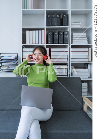 Woman sitting comfortably on a couch with headphones, relaxing to music while using a computer in a cozy setting Woman sitting comfortably on a couch with headphones, relaxing to music while using a computer in a cozy setting 123578578