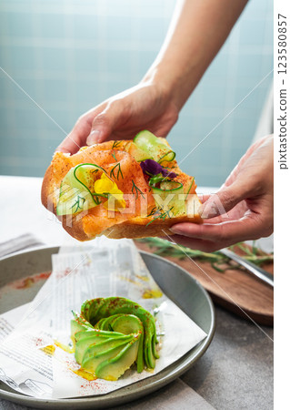 In the cafe setting, hold the cucumber, avocado, and smoked salmon sandwich with both hands In the cafe setting, hold the cucumber, avocado, and smoked salmon sandwich with both hands 123580857