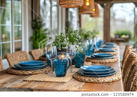Elegant outdoor dining table set with blue plates, glasses, and wicker chairs in a cozy patio with greenery 123580974