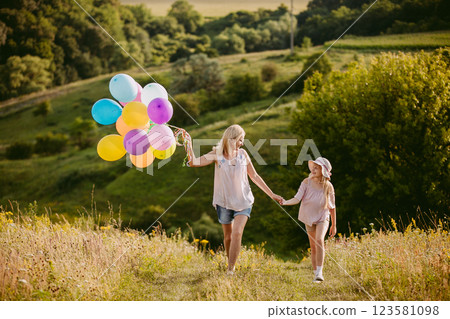 Happy girl with colorful balloons holding hands with a smiling middle-aged woman, walking together in a sunny meadow surrounded by greenery. Happy girl with colorful balloons holding hands with a smiling middle-aged woman, walking together in a sunny meadow surrounded by greenery. 123581098