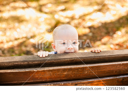 Almost bald baby boy peeking out from behind a wooden bench in the autumn park Almost bald baby boy peeking out from behind a wooden bench in the autumn park 123582560