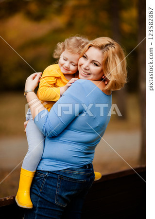 Loving mother holding toddler girl with curly hair in yellow shirt in autumn park 123582867