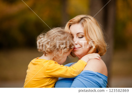 Loving mother holding toddler girl with curly hair in yellow shirt in autumn park 123582869