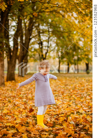 Toddler girl in purple dress, white tights, and yellow rubber boots playing happily with leaves in autumn park 123582968