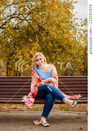 Plus-size blonde woman in jeans and blue shirt sitting on a bench with a blanket on shoulders in autumn park 123582978