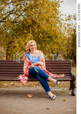 Plus-size blonde woman in jeans and blue shirt sitting on a bench with a blanket on shoulders in autumn park Plus-size blonde woman in jeans and blue shirt sitting on a bench with a blanket on shoulders in autumn park 123582980
