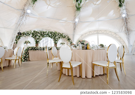 Elegant wedding reception hall featuring round tables, white and gold chairs, and floral arrangements Elegant wedding reception hall featuring round tables, white and gold chairs, and floral arrangements 123583098