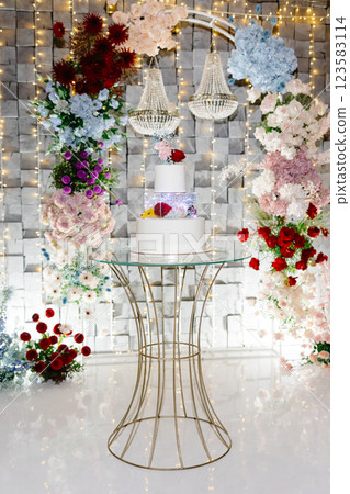 Elegant white wedding cake displayed on a golden stand, surrounded by colorful flower arrangements and chandeliers Elegant white wedding cake displayed on a golden stand, surrounded by colorful flower arrangements and chandeliers 123583114