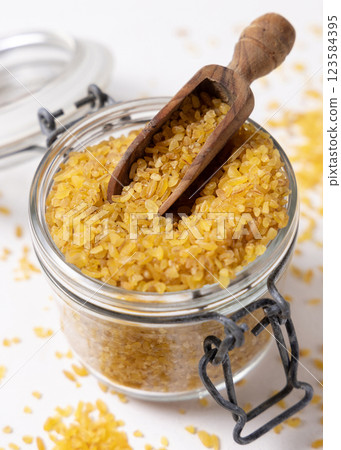 Glass jar of raw dry uncooked yellow bulgur grain with a wooden scoop on napkin closeup Glass jar of raw dry uncooked yellow bulgur grain with a wooden scoop on napkin closeup 123584395
