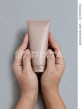 Hands holding light brown squeeze cream tube over grey top view, cosmetic mockup, copy space 123584412
