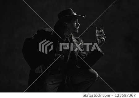 Black and white portrait of man admiring whiskey glass, dressed in leather coat and hat against dark, dramatic background. 123587367