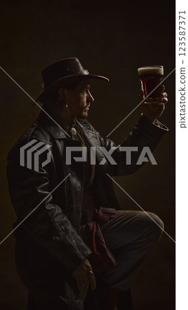 Cowboy raising pint of beer, admiring rich color and foam, dressed in classic western outfit with leather coat and hat 123587371