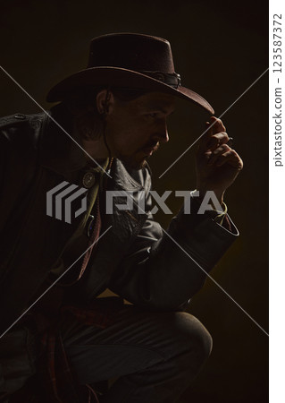 Portrait of brutal thoughtful man, cowboy dressed in leather coat and hat exuding silent strength and wisdom against dark background. 123587372