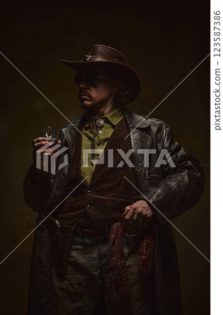 Dark and moody portrait of man, cowboy in leather coat and vest, gripping whiskey glass, standing in confident stance. 123587386