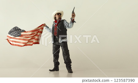 Powerful cowboy in white hat and leather vest, pointing revolver forward, gripping American flag, wearing sunglasses and boots. Powerful cowboy in white hat and leather vest, pointing revolver forward, gripping American flag, wearing sunglasses and boots. 123587390