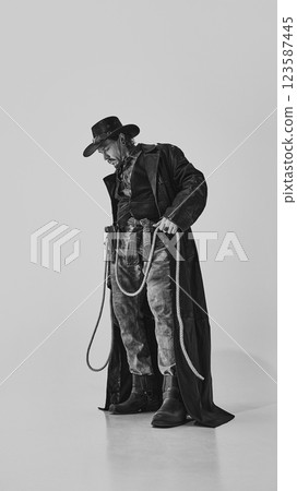 Full-length portrait of brutal man in jeans, leather coat, boots and hat, standing with lasso in hand. Calm confidence. 123587445