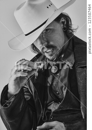 Black and white portrait of handsome man dressed in leather coat and white fedora hat, gripping cigarette with intense gaze. 123587464