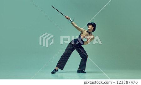 Charismatic man in vintage corset suit and bow tie elegantly balancing with cane against muted green studio background. 123587470
