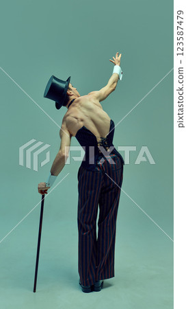 Back view of poised artist in structured corset and pinstripe trousers extending arm with refined elegance against muted green studio background. 123587479