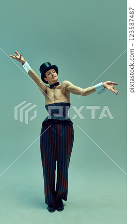 Enigmatic figure in structured corset suit and sleek hat raising cane dramatically against muted green studio background. Enigmatic figure in structured corset suit and sleek hat raising cane dramatically against muted green studio background. 123587487