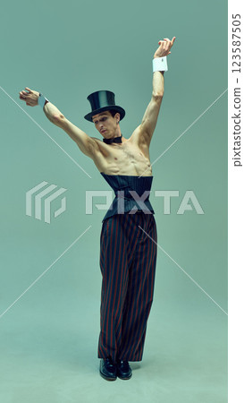 Dynamic photo of performer in high-waisted corset and classic hat extending arms dramatically against muted green studio background. 123587505