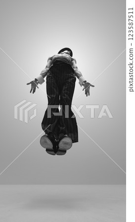 Black and white photo of elegant man, performer in striped trousers and hat floating mid-air with arms extended against white studio background. 123587511