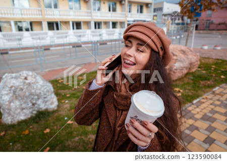 Top view of young pretty Caucasian happy woman in a coat and hat holds cup of coffee call on smartphone in city street. Lifestyle and city life Top view of young pretty Caucasian happy woman in a coat and hat holds cup of coffee call on smartphone in city street. Lifestyle and city life 123590084