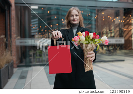 Happy smiling woman wearing black long trench coat, holds a red spring gift package bag and spring flowers, walking in street of European city Happy smiling woman wearing black long trench coat, holds a red spring gift package bag and spring flowers, walking in street of European city 123590519