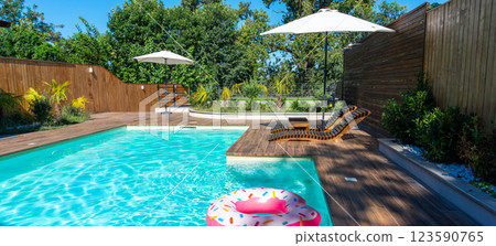 Modern backyard swimming pool with turquoise water, wooden deck, umbrellas, lounge chairs, and pink inflatable ring 123590765